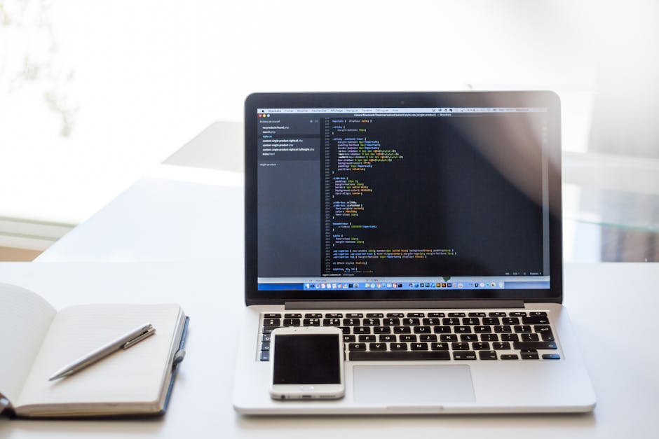 A black-and-white photograph of a workspace featuring an open laptop displaying lines of code on its screen, placed on a dark wooden desk. To the right of the laptop, there is a white notebook with a 