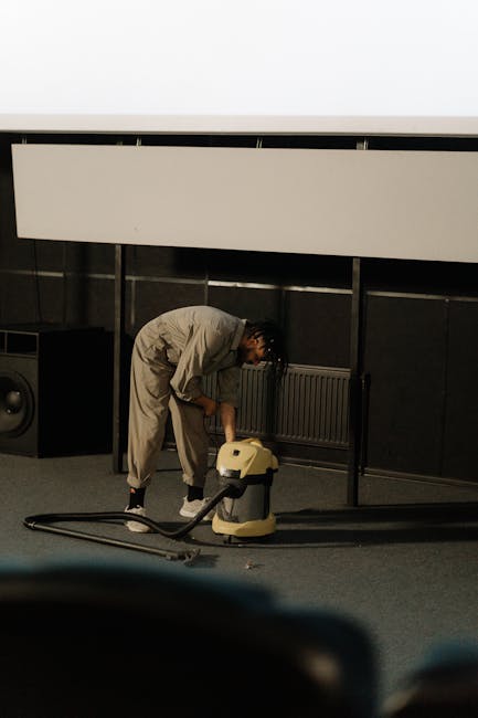 A professional cleaner dressed in a white protective suit, mask, gloves, and shoe covers is performing deep cleaning on a beige carpet in a well-lit living room. The cleaner is using a steam or extrac
